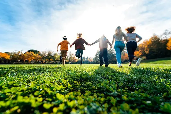 A group of people holding hands run across a grassy field towards the sun. The bright sunlight creates a vibrant, lively atmosphere, with autumn trees in the background adding a touch of color to the scene.