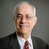 A man wearing glasses and a suit with a patterned red tie, smiling at the camera against a gray background.