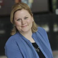 Woman with blonde hair wearing a blue blouse and necklace, smiling at the camera with a dark background.