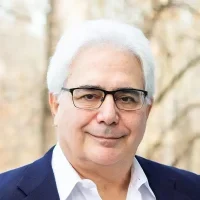 Older man with white hair, glasses, and a suit jacket, smiling outdoors with a blurry background.