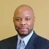 A man in a suit and tie smiles slightly in front of a plain beige background.