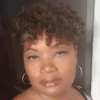 Woman with short curly hair, wearing hoop earrings, looking at the camera with a neutral expression.