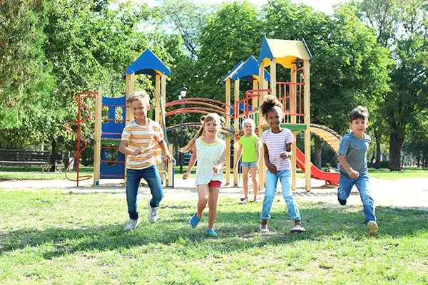 Five children running and smiling in front of colorful playground equipment in a sunny park.