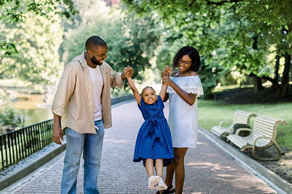 A smiling couple swings a happy little girl between them while walking in a sunny, tree-lined park.
