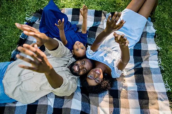 A joyful family of three lies on a blanket outdoors, smiling and reaching their arms toward the camera.