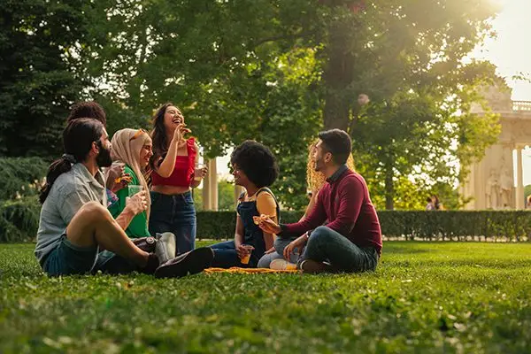 Six friends sitting on grass in a park, laughing and enjoying food together on a sunny day.