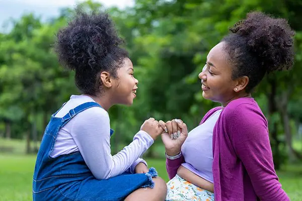 A mother and daughter sit outside, smiling and linking pinkies, with trees in the background.