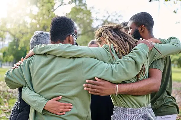 Five people standing outdoors in a circle, arms around each other, sharing a group hug.