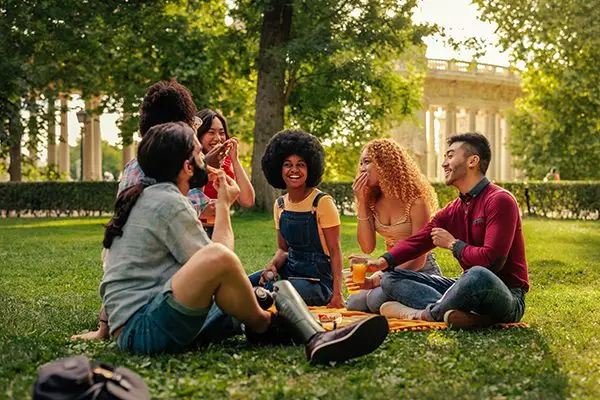 A group of friends sit on grass in a park, laughing and enjoying drinks together on a sunny day.