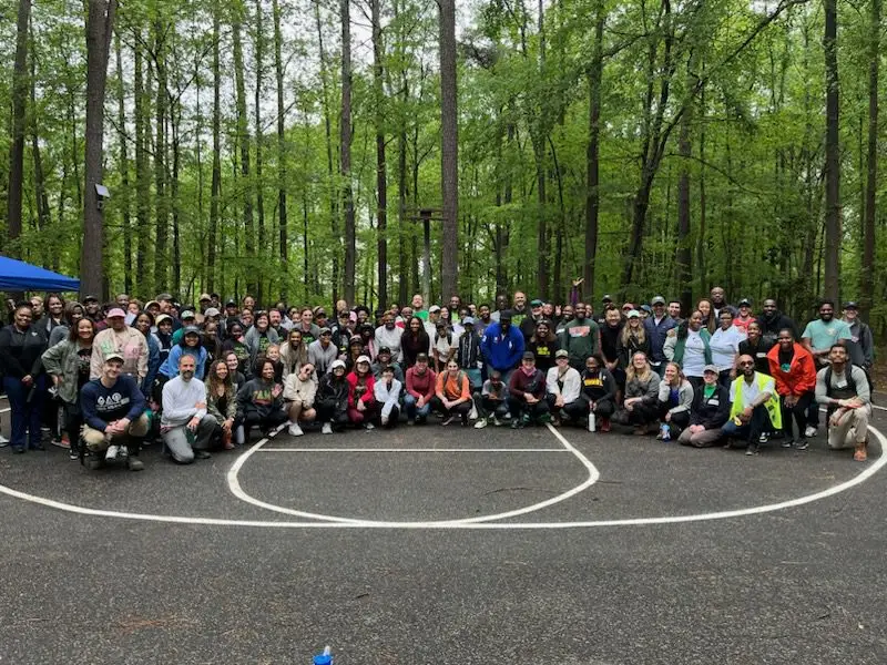 Large group of people posing together on an outdoor basketball court surrounded by tall trees in a forest.