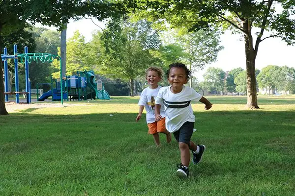 Two young children run and smile in a grassy park built with the help of generous donation, playground equipment behind them.