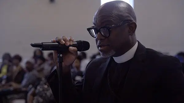 A man in glasses speaks into a microphone at an indoor gathering, discussing upcoming events with people seated in the background.