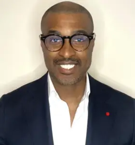 Smiling man in glasses, dark blazer, and white shirt, embodying community advocacy against a plain light background.