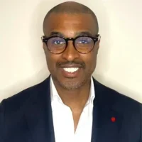 Smiling man in glasses, dark blazer, and white shirt, embodying community advocacy against a plain light background.