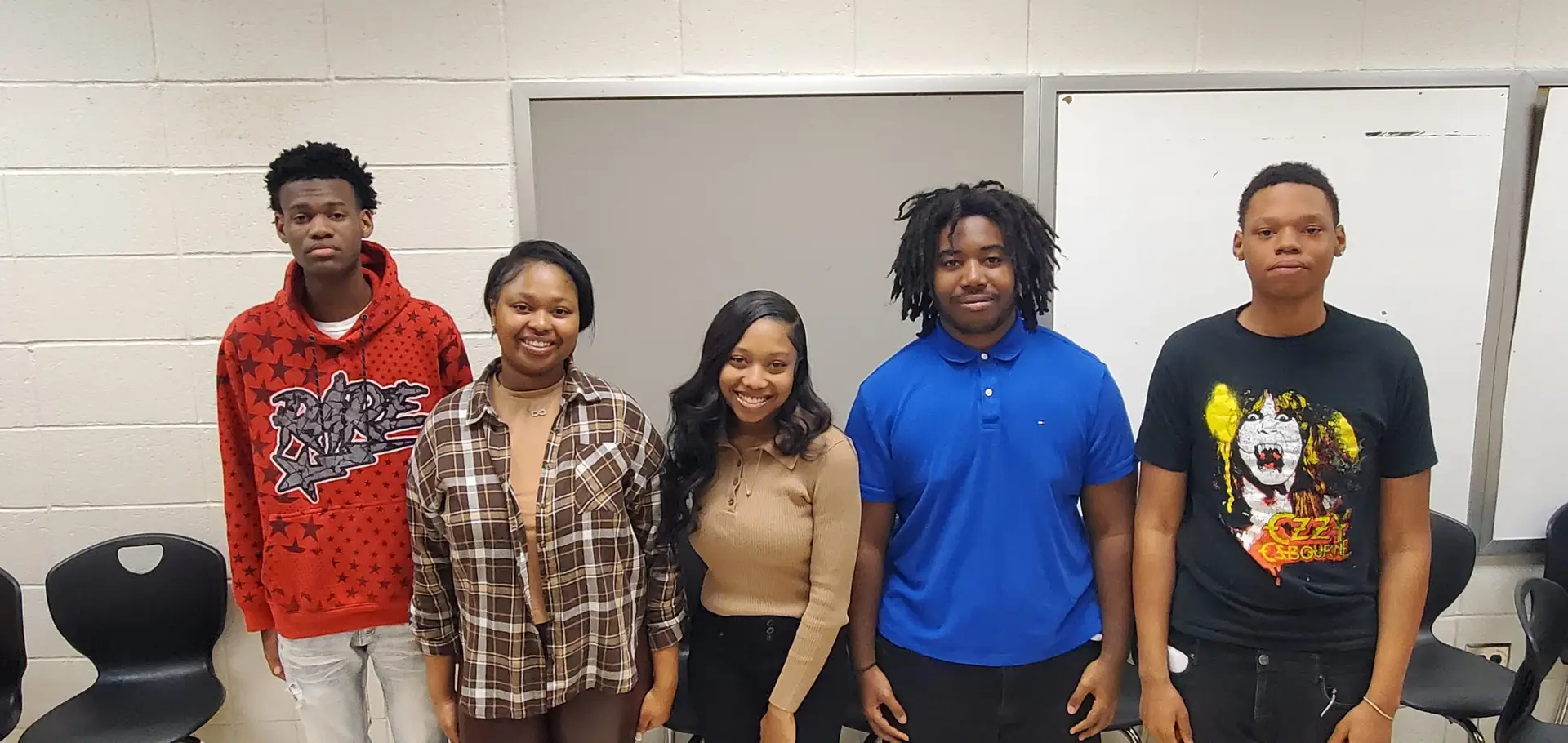 Five young people standing indoors, smiling, in front of a white and gray classroom wall.