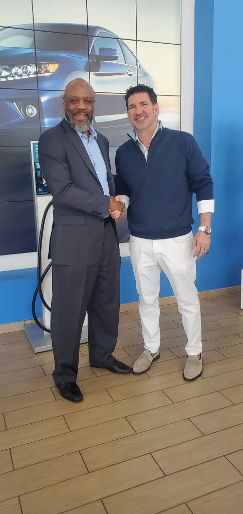 Two men shaking hands and smiling in front of a Lou Sobh Automotive Group car display at a dealership.