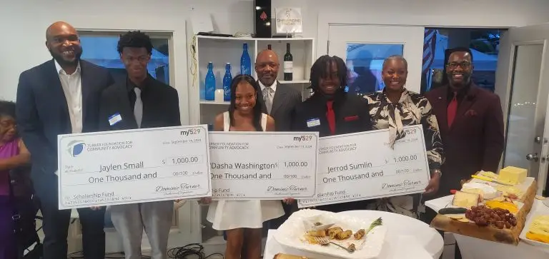 Three students hold large scholarship checks, posing and smiling with four adults at a celebratory event.