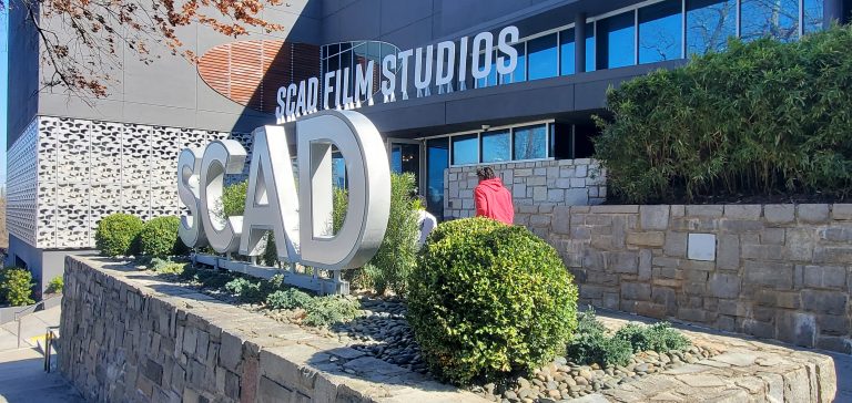 Large SCAD sign and SCAD Film Studios building exterior with two people walking nearby, surrounded by greenery.