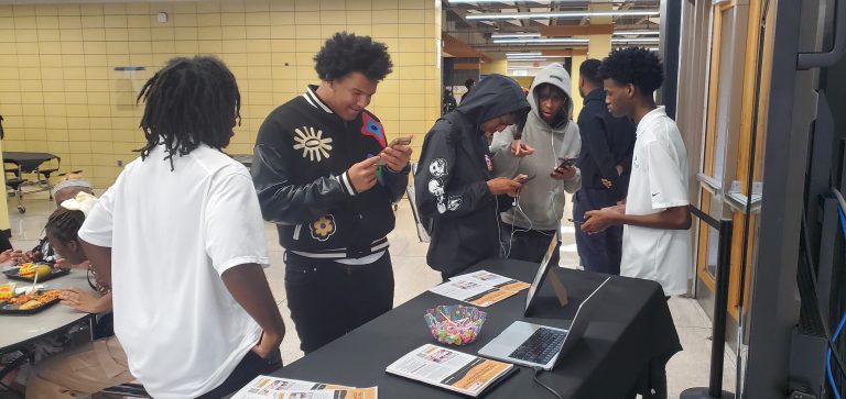 Year Two Participants stand around a table with flyers, a laptop, and candy in a school hallway.