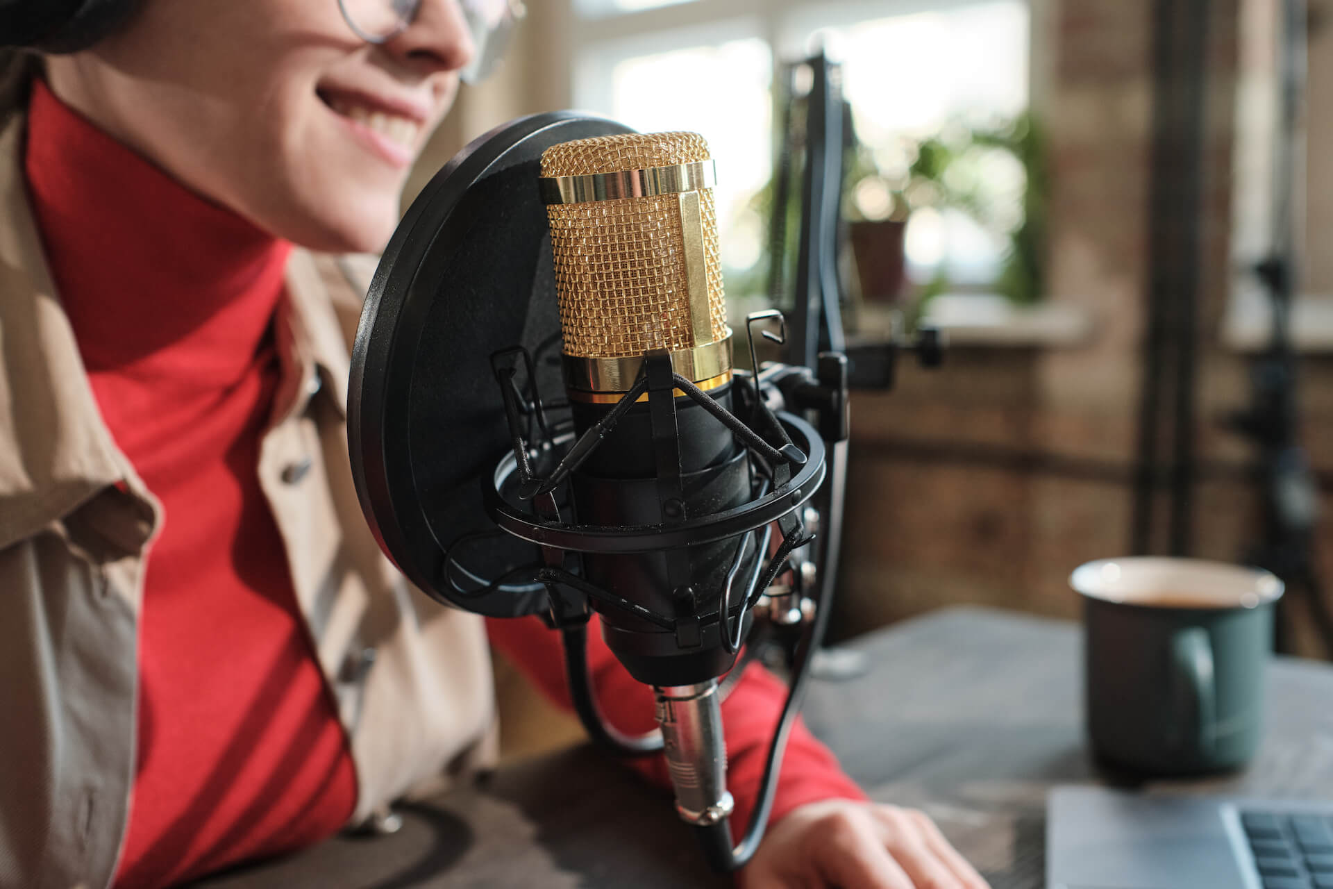 Person smiling and speaking into a microphone with a pop filter, recording at a desk with a mug nearby.