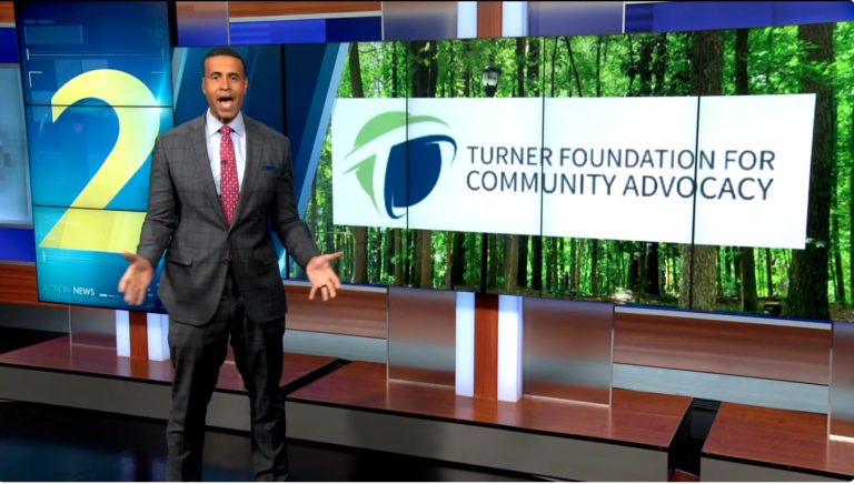 News anchor stands beside a screen displaying Turner Foundation for Community Advocacy logo and forest background.