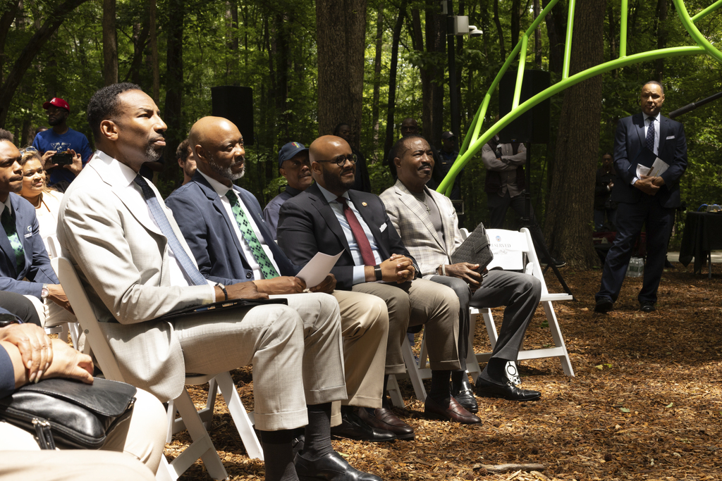 A group of men in suits sit outside on white chairs during a formal event in a wooded area.