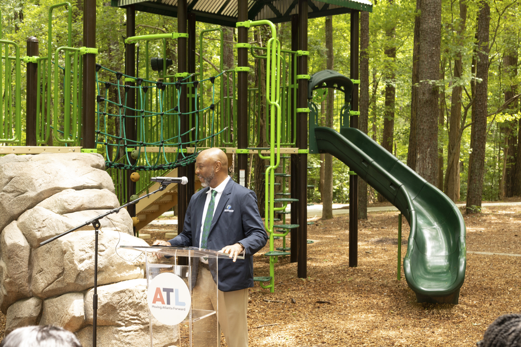 A man speaks at a podium in front of a playground with slides and climbing equipment in a wooded park.