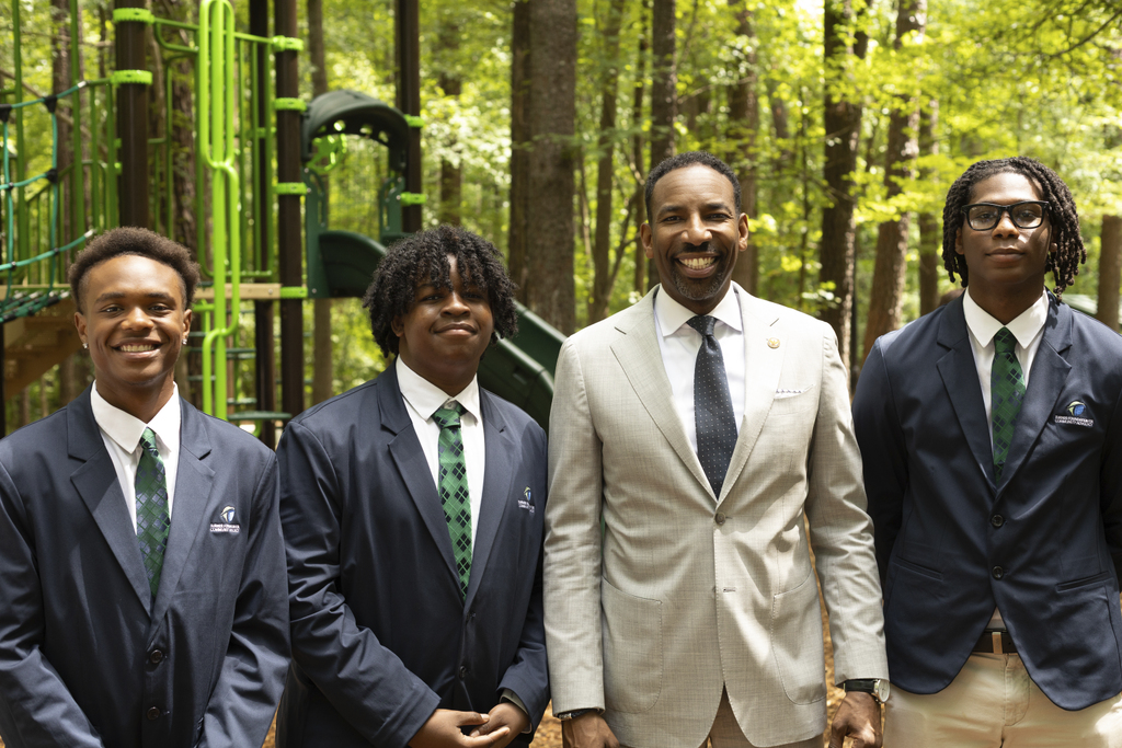 Four men in suits stand and smile in front of a green playground in a wooded outdoor setting.