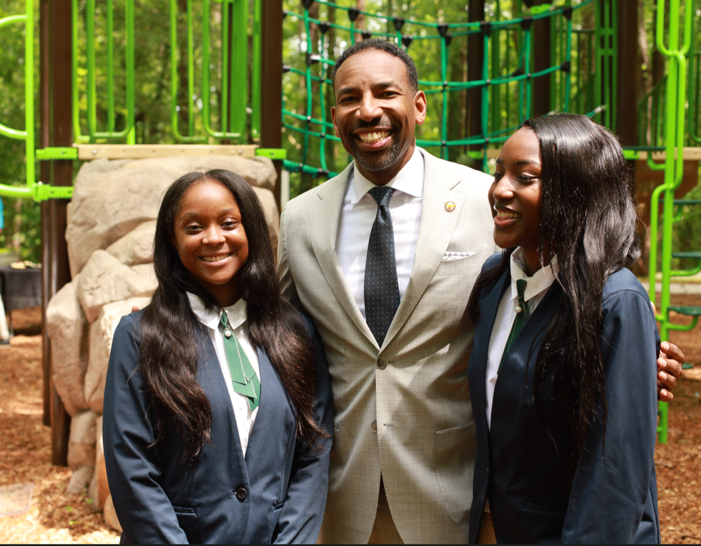 A man in a suit stands smiling with two young women in uniforms at a playground outdoors.