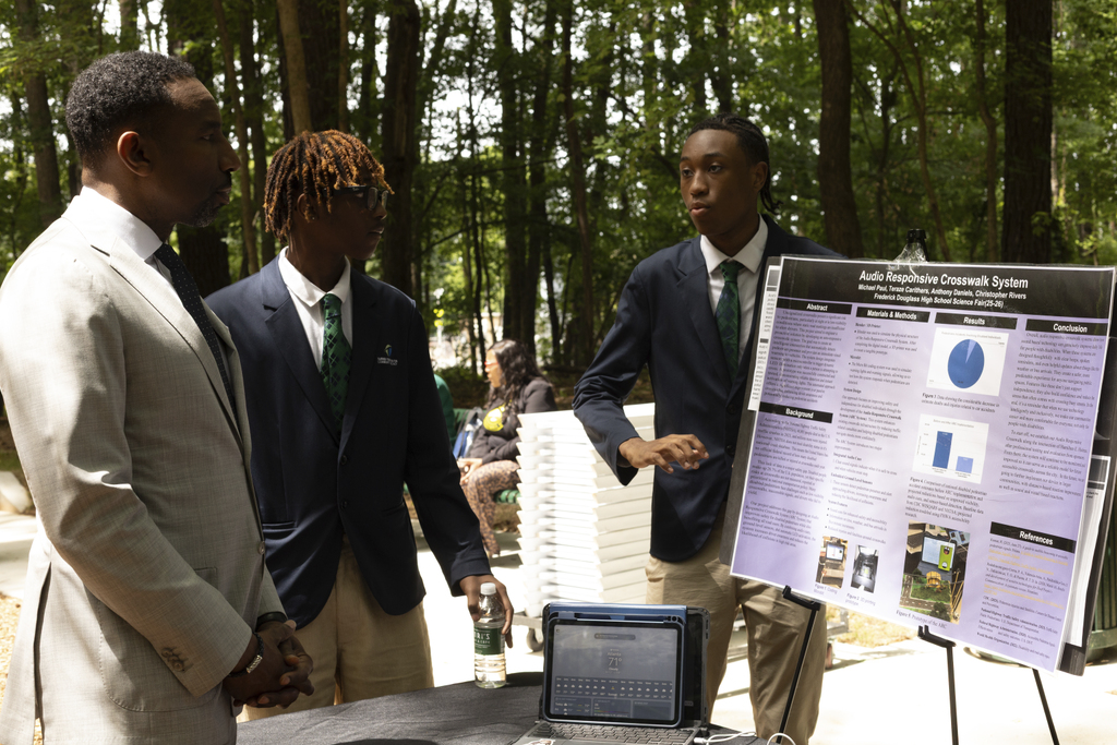 Three young men in suits discuss a science project with a display board and laptop outdoors amid trees.