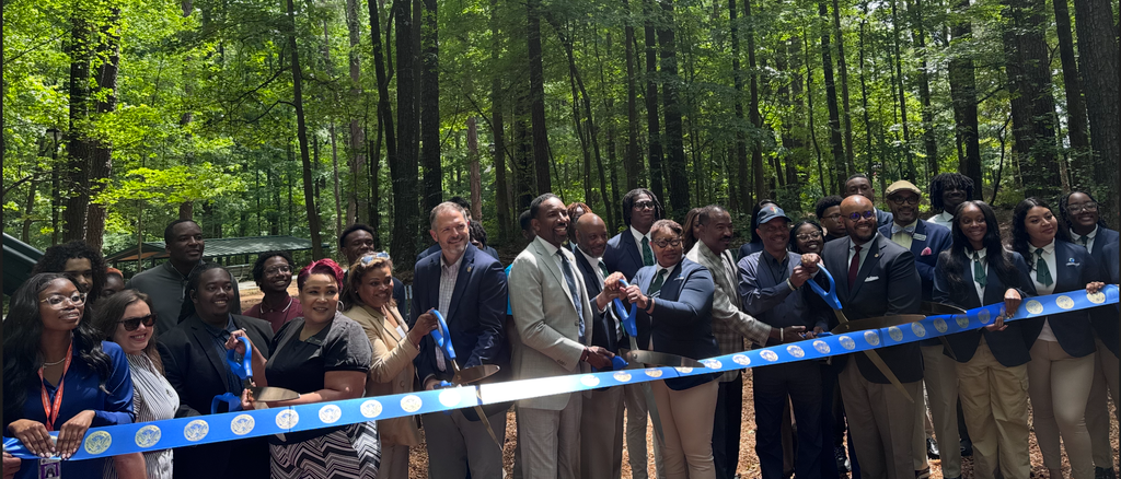 A group of people stand in a forest, smiling and cutting a long blue ribbon at a ribbon-cutting ceremony.