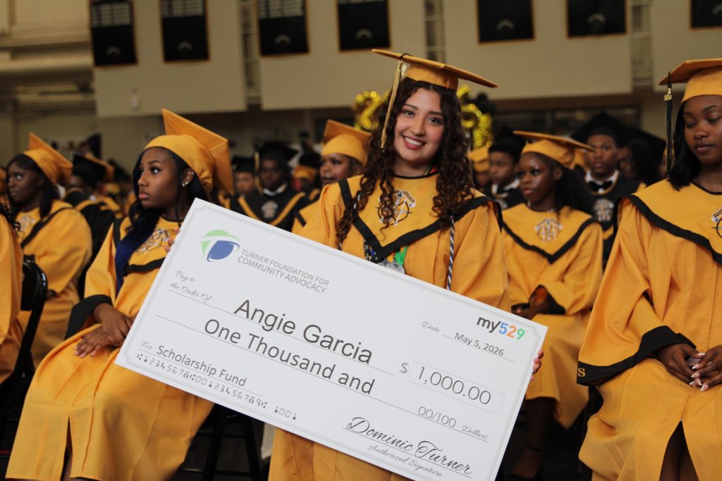 A graduate in a yellow cap and gown holds a 1,000 scholarship check at a graduation ceremony.