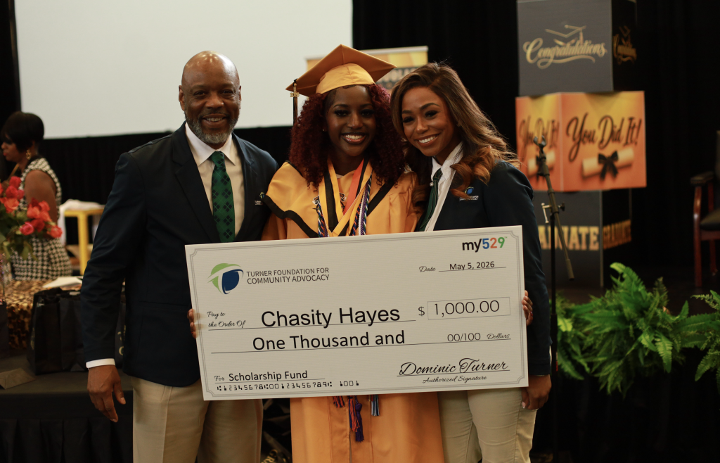 Three people smile as a graduate holds a large 1,000 scholarship check at a graduation event.