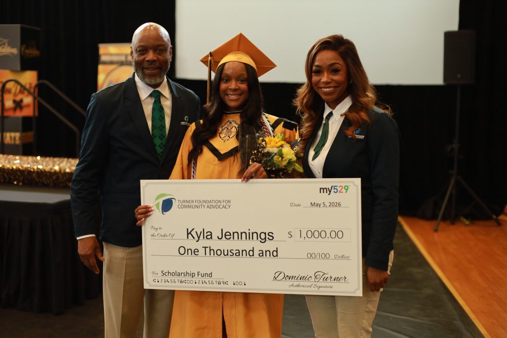 A graduate in a gold cap and gown holds a 1,000 scholarship check, standing with two smiling adults.