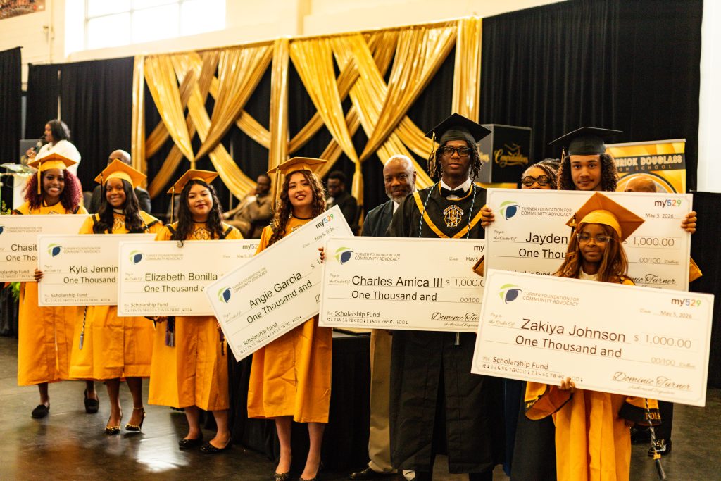 Seven graduates in gold caps and gowns hold large scholarship checks and smile at a graduation ceremony.