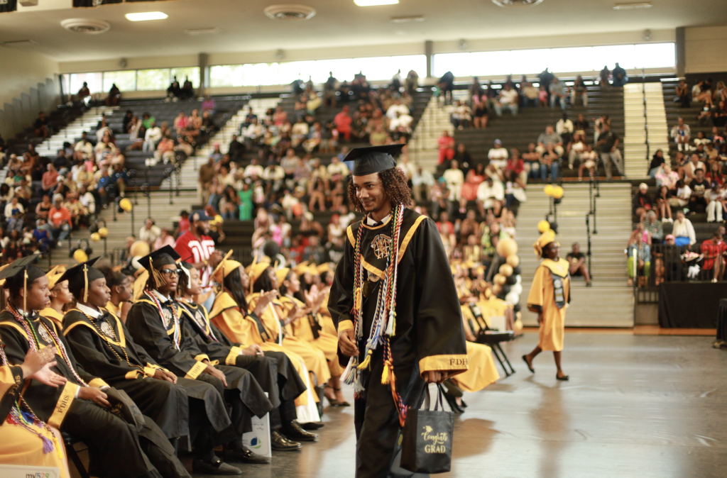 A graduate in cap and gown walks in a gymnasium as others sit, with a crowd in the bleachers behind.
