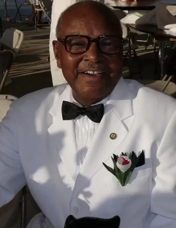 Smiling man in a white tuxedo with a black bow tie and glasses, sitting outdoors at an event.