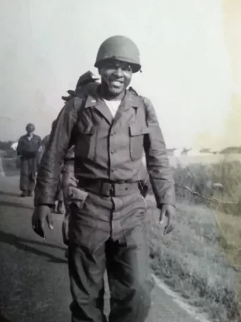 Black-and-white photo of a smiling soldier in uniform and helmet walking down a road, others in background.