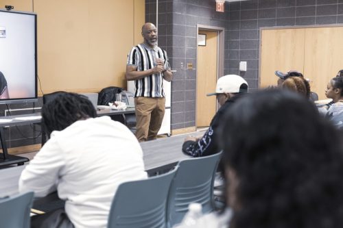 A man stands and speaks to a seated group in a classroom setting with a screen and tables.