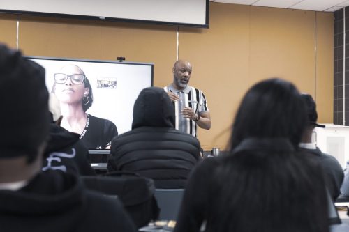 A man speaks to an audience in a classroom, with a woman on a video call displayed behind him.