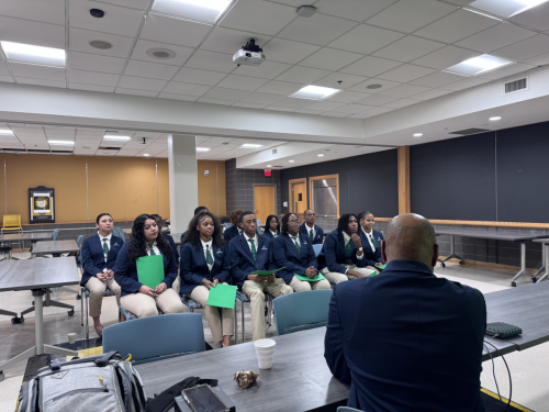 A group of students in blazers sit facing an instructor in a classroom, holding green folders.