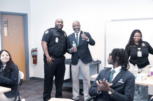 Two men, one in a police uniform and one in a suit, smile and pose in a classroom with students.