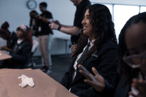 A student in a blazer sits at a table with gloves, smiling, as others work in the background.