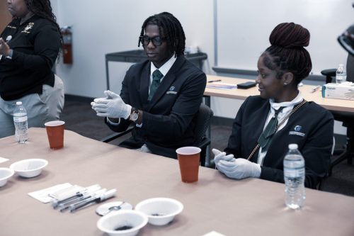Two people in suits and gloves sit at a table with cups, bowls, water bottles, and small tools in a classroom.