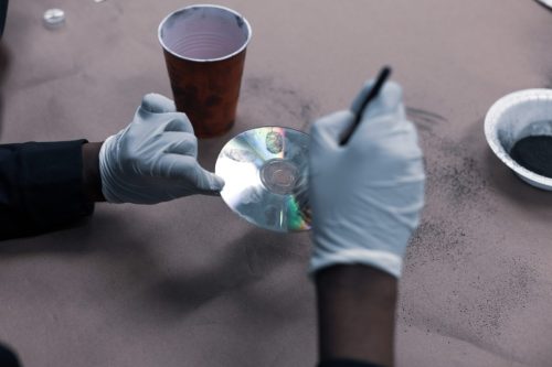 Gloved hands holding a CD and using a tool, with a cup and a plate of black powder on a table.