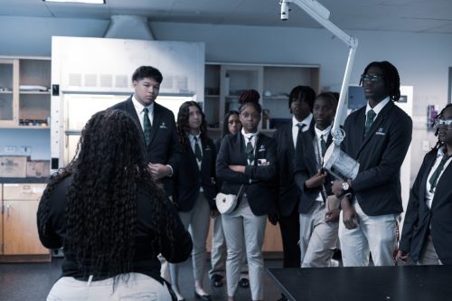 Students in uniforms listen to a teacher in a science classroom with lab equipment and cabinets in the background.
