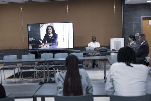 People in a meeting room watch a large screen showing a person speaking in a virtual call.