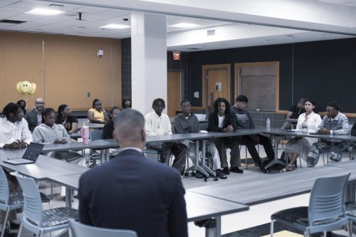 A group of people sit around tables in a classroom, listening to a speaker at the front of the room.