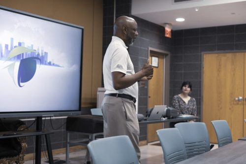 A man gives a presentation in a conference room, while a woman sits at a table with a laptop.