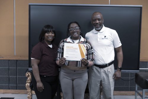 Three people smile as the person in the center holds a certificate in front of a large monitor.